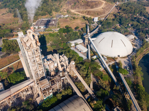 Aerial view of an industrial cement plant featuring a large white dome structure, tall smoke stacks with emissions, and extensive conveyor belt systems surrounded by lush greenery in Indonesia.