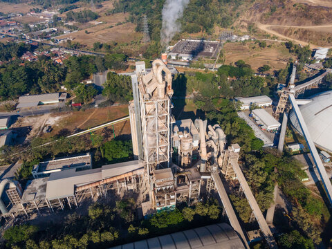 Aerial view of a large industrial cement factory with tall processing towers, pipes, and a smoke stack emitting steam, set within a rural landscape in Indonesia.