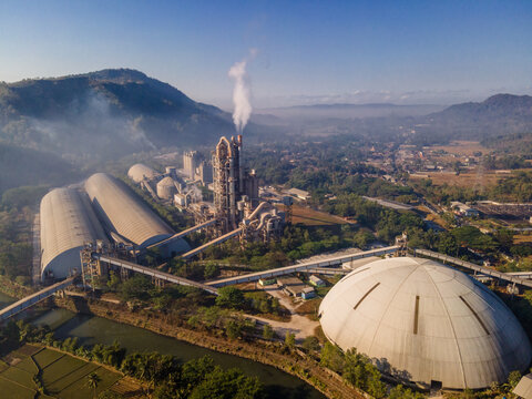 Aerial view of a large industrial cement factory complex with a tall chimney emitting smoke, large storage domes, and conveyor belts, situated amidst lush green hills in Indonesia.