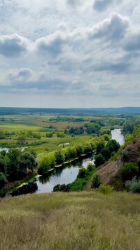 Panoramic stock video of a calm winding river flowing through a green countryside valley with lush trees and fields under a cloudy sky.
