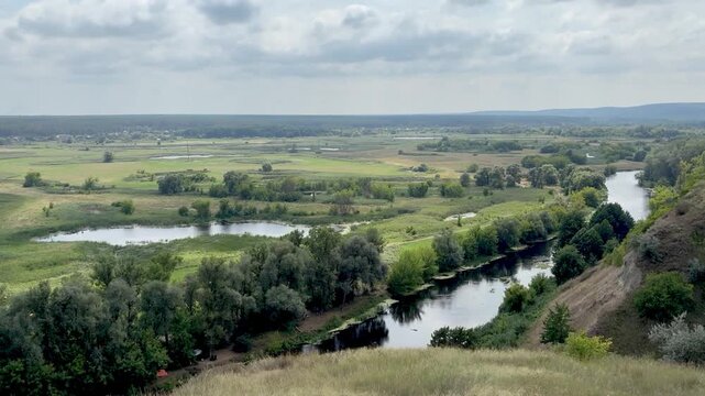 Panoramic stock video of a calm winding river flowing through a green countryside valley with lush trees and fields under a cloudy sky.
