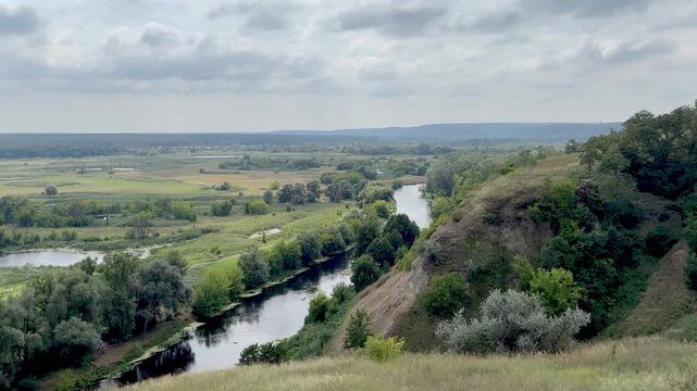 Panoramic stock video of a calm winding river flowing through a green countryside valley with lush trees and fields under a cloudy sky.
