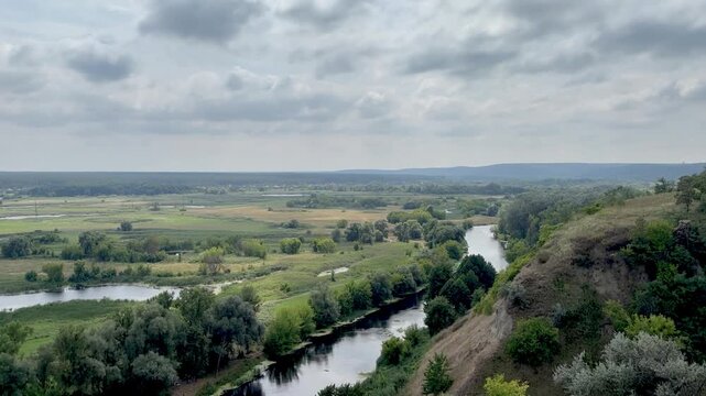 Panoramic stock video of a calm winding river flowing through a green countryside valley with lush trees and fields under a cloudy sky.
