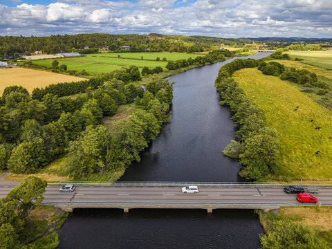 Aerial view of the Viskan river flowing under a bridge with cars driving across, surrounded by lush green fields, trees, and agricultural land in Askloster, Halland County, Sweden.