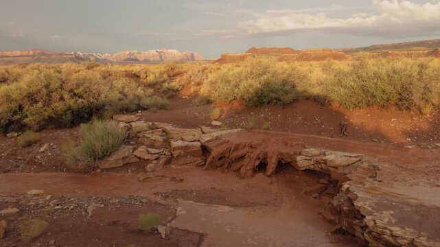 Red muddy water pours over a sandstone ledge in a desert ravine after a summer thunderstorm has past while shadows stretch towards the distant mountains of Zion NP Utah USA.