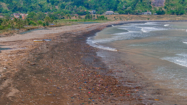 Aerial view of plastic waste and debris scattered across a sandy beach, highlighting environmental pollution with waves washing onto the shore and green hills in the background in Indonesia.