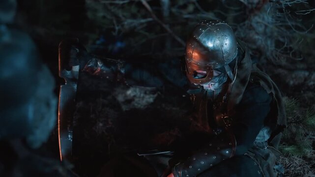 Dark wet forest at night shows two masked figures in tense close encounter, pale mask dominating foreground while helmeted figure crouches behind amid mud, foliage, shadows, bloodstains, and uneasy