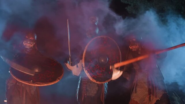 Three armored figures march forward in tight formation with round shields held ahead and long weapons lifted, sparks, ember glow, and swirling smoke heightening tense battle mood, with shield, armor