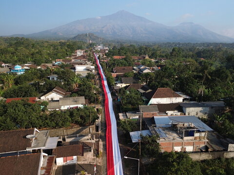 Aerial view of a long red and white Indonesian flag draped along a village street, surrounded by houses and greenery, with a mountain in the background in Wonosari, East Java, Indonesia.