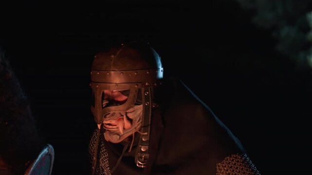 Helmeted medieval warrior in chainmail and worn metal armor swings wooden pole weapon with body turned, intense gaze visible through helmet slit, and warm firelight revealing soot, scratches, and