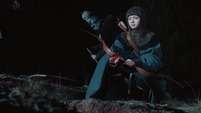 Young woman crouched outdoors at night releases arrow from compound bow, showing focused posture, extended arms, slight body rotation, and tense motion after shot in cool rim light