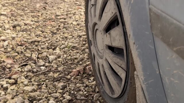 Dirty car wheel parked on rocky gravel, mud-splattered hubcap, compressed tire sidewall, and worn wheel arch shown from low angle with rough texture and overcast light