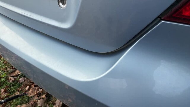 Close-up rear trunk opening on light blue car revealing bumper edge, rubber seal, scuffed threshold, seam rust, tail light contrast, and detailed body panel gap