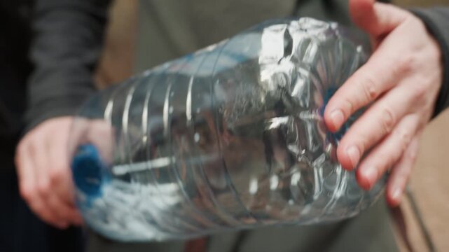 Tight crop showing two hands gripping and tilting large clear plastic water bottle, creating visible liquid motion, internal ripples, and angled flow in outdoor daylight with practical, cooperative