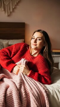Young woman in a red sweater relaxing on a bed with a chunky pink blanket in a cozy bedroom, smiling in warm natural light