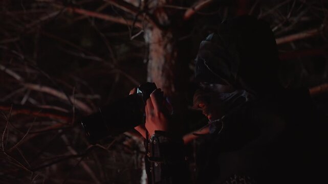 Nighttime close view of hooded person steadying rifle against shoulder while peering along its length, with concealed face, warm red sidelight, dark pine trees, and tense watchful mood