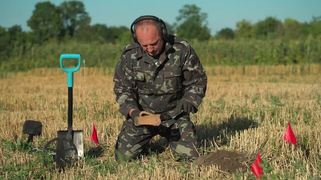 Male soldier in camouflage uniform kneels in a field, using a small tool to examine soil while surrounded by red flags marking potential excavation sites and a shovel resting nearby