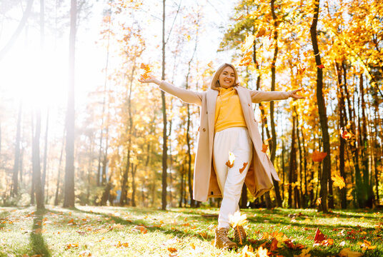 Smiling woman in coat sits in clearing in park surrounded by fallen leaves at sunset. Woman tosses bright yellow leaves into the air, enjoying the outdoors. Concepts of fun and enjoyment.