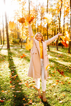 Smiling woman in coat sits in clearing in park surrounded by fallen leaves at sunset. Woman tosses bright yellow leaves into the air, enjoying the outdoors. Concepts of fun and enjoyment.