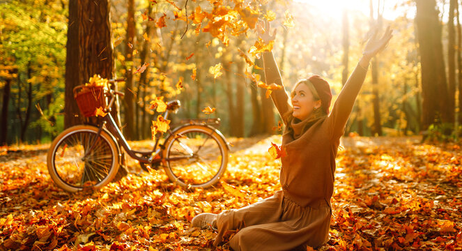Woman is having fun in an autumn park, overlooking bicycle at sunset. Young woman throws bright yellow leaves while walking through sunny park. Concepts of relaxation, walking, and seasonality.