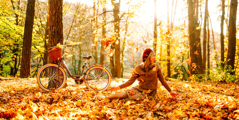 Fototapeta premium Woman is having fun in an autumn park, overlooking bicycle at sunset. Young woman throws bright yellow leaves while walking through sunny park. Concepts of relaxation, walking, and seasonality.