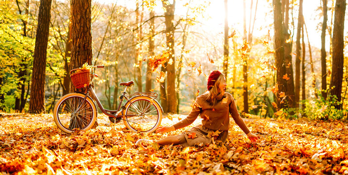 Woman is having fun in an autumn park, overlooking bicycle at sunset. Young woman throws bright yellow leaves while walking through sunny park. Concepts of relaxation, walking, and seasonality.