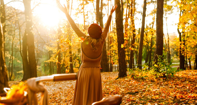 Woman is having fun in an autumn park, overlooking bicycle at sunset. Young woman throws bright yellow leaves while walking through sunny park. Concepts of relaxation, walking, and seasonality.