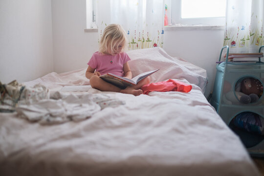 Little Girl Reading and Turning Pages of a Book on a Bed