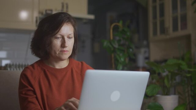 The woman works intently on her laptop, with a focused expression. The warm lighting and green plants create a calm atmosphere in this home office setting.