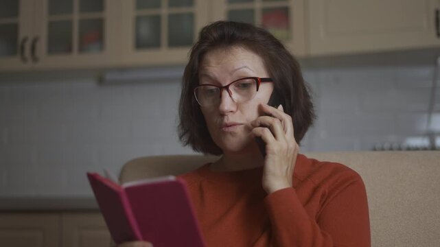 A woman sits on a sofa, holding a notebook while talking on a telefon. The woman appears focused on her telefon, glancing at the notebook for reference.