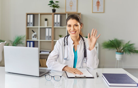 Friendly smiling female doctor looking at camera having online video call or conference waving hand greeting her colleague or patient. Telemedicine and patient consultation online concept.