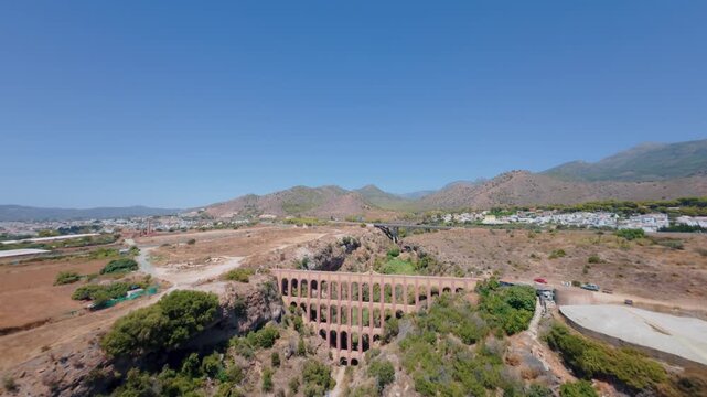 FPV-Drohnenflug &uuml;ber ein spanisches Tal mit historischen Aqu&auml;dukt und zwei Br&uuml;cken nahe M&aacute;laga, Berglandschaft und Dorf im Hintergrund