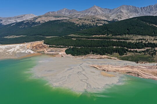 Aerial view of Mayflower Tailings Pond with sediment patterns, surrounded by evergreen forests and rugged Rocky Mountain peaks in Summit County, Colorado, United States.