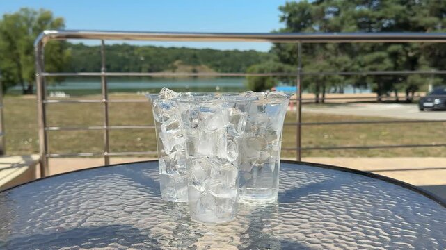 Three plastic cups packed with crystal clear ice cubes sparkle in bright sunlight on a glass table, creating a refreshing summer scene for outdoor dining and relaxation. Close up.