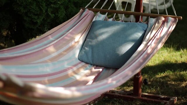 An empty striped, colorful hammock with a blue pillow that gently sways from one side to the other in the summer breeze.