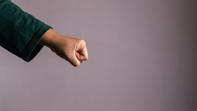 A single human arm extends a tightly clenched fist toward the right side against a solid grey studio backdrop. The unrecognizable Asian male wears a dark green shirt, projecting a