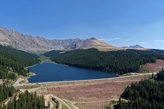 Aerial view of Clinton Gulch Dam Reservoir surrounded by dense pine forests and rugged mountain slopes under a clear blue sky in Summit County, Colorado, United States.