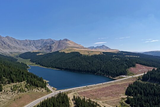 Aerial view of Clinton Gulch Dam Reservoir surrounded by dense pine forests and rugged mountain peaks under a clear blue sky in Summit County, Colorado, United States.