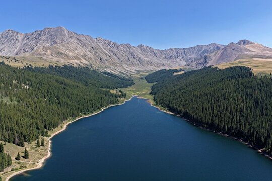 Aerial view of Clinton Gulch Dam Reservoir surrounded by dense pine forests and rugged mountain peaks under a clear blue sky in Summit County, Colorado, United States.