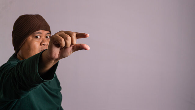 A focused Southeast Asian male extends an arm toward the camera to make a pinching gesture with the fingers against a plain grey studio backdrop. The adult wears a brown beanie and