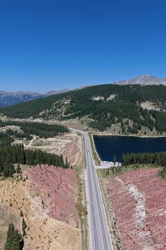 Aerial view of Clinton Gulch Dam Reservoir with a highway running alongside, surrounded by pine forests and rocky slopes under a clear blue sky in Summit County, Colorado, United States.
