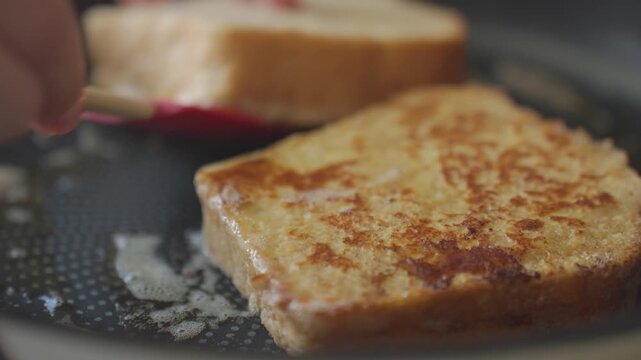 Hand flips French toast with red spatula in hot frying pan as bread browns in butter. Concept of breakfast cooking and pan frying
