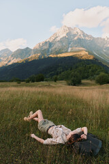 Fototapeta premium A woman rests in a sunlit meadow with a backpack beside her, mountains rising in the distance. A calm travel scene conveying rest, exploration, and connection with nature.