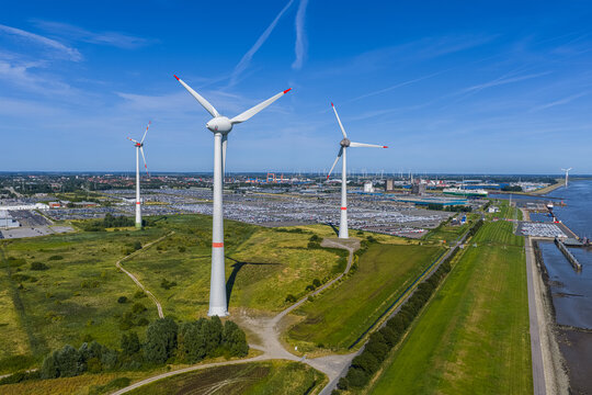 Aerial view of wind turbines at the Volkswagenwerk Emden plant with vast car storage lots and green fields in Emden, Niedersachsen, Germany.
