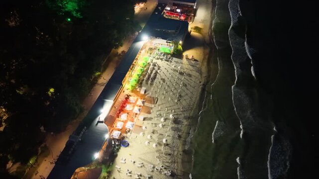 Top-down aerial night view of an illuminated seaside beach with restaurants, rows of sun loungers and umbrellas beside dark ocean waves, showing vibrant coastal nightlife and tourism.