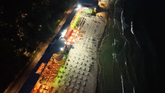 Top-down aerial night view of an illuminated seaside beach with restaurants, rows of sun loungers and umbrellas beside dark ocean waves, showing vibrant coastal nightlife and tourism.