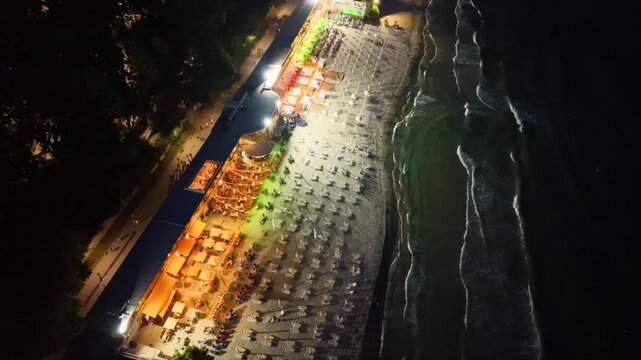 Top-down aerial night view of an illuminated seaside beach with restaurants, rows of sun loungers and umbrellas beside dark ocean waves, showing vibrant coastal nightlife and tourism.