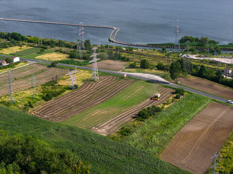 Aerial view of a combine harvester working in fields near power lines and a lake, with rural landscape and infrastructure in Konin, Wojewodztwo wielkopolskie, Poland.