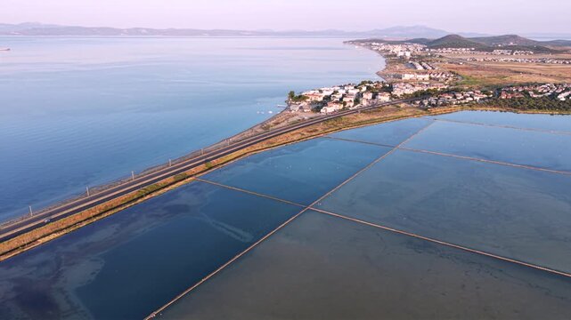 Aerial view of coastal salt evaporation ponds beside a highway and calm sea, showing pink and blue saline pools, shoreline infrastructure, farmland and distant mountains at sunset.