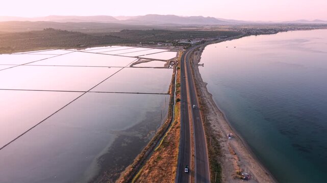 Aerial view of coastal salt evaporation ponds beside a highway and calm sea, showing pink and blue saline pools, shoreline infrastructure, farmland and distant mountains at sunset.